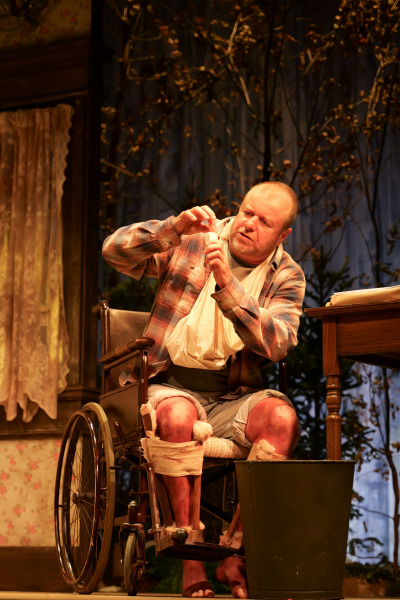 A tense scene from Misery features a disheveled man in a wheelchair examining a small bottle of pills under warm stage lighting. His legs are heavily bandaged with visible bruises and blood stains, and his arm is in a makeshift sling. He wears a plaid shirt and grey shorts, sitting beside a wooden desk with a green trash bin in front. Behind him, lace curtains and bare branches add to the unsettling, rustic atmosphere.