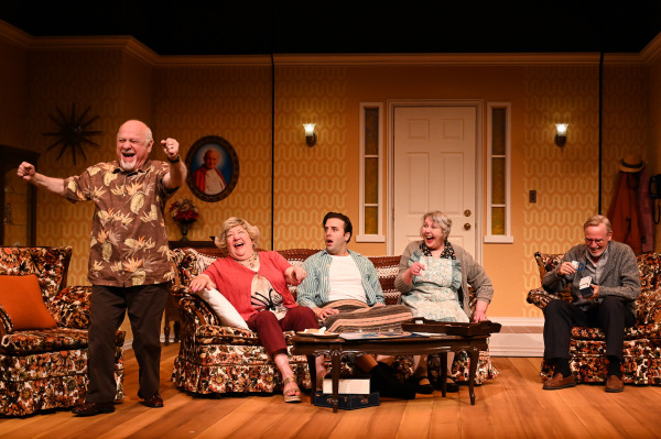 In this image from Over The River and Through The Woods, a joyful theatre scene set in a cozy living room. An older man stands triumphantly with his fists raised, while two older women, a young man, and another older man sit on floral couches, laughing and reacting. One woman clutches a gift, and the man on the right inspects a pair of socks. The warm, vintage set features patterned wallpaper, a family portrait, and retro furniture, creating a lively, heartwarming atmosphere.