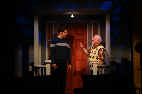 In this image from Over The River and Through The Woods, a nighttime theatre scene on a front porch. A young man in a dark sweater with white stripes listens intently as an older man in a patterned vest and tropical shirt points and smiles warmly. The porch set features a red door, white railing, and soft blue lighting, creating an intimate, heartfelt atmosphere.