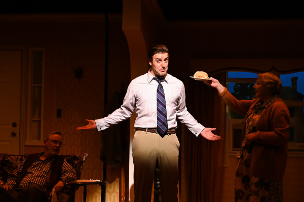 In this image from Over The River and Through The Woods, a comedic moment on stage in a cozy living room set. A man in a striped dress shirt, tie, and khakis stands with his arms outstretched and an exaggerated, puzzled expression. A woman in a warm-toned sweater offers him a plate with a sandwich. In the background, an older man sits in a floral armchair watching the interaction. The scene is warmly lit, emphasizing the playful family atmosphere.