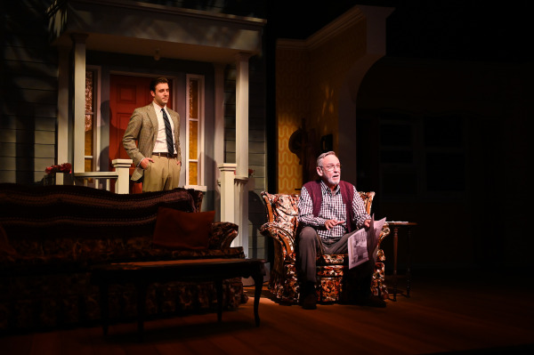 In this image from Over The River and Through The Woods, a theatre scene set in a cozy living room and front porch. An older man sits in a floral armchair holding a newspaper, wearing a checkered shirt and maroon sweater vest, mid-conversation. Behind him, a younger man in a tan blazer, dress shirt, and tie stands on the porch with his hands in his pockets, listening. The stage is warmly lit, highlighting the homey set with vintage furniture and patterned wallpaper.