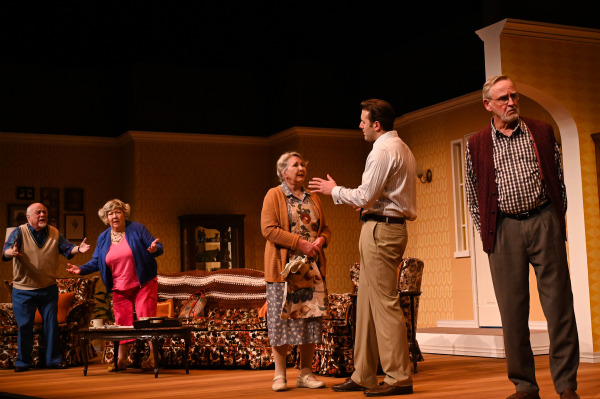 In this image from Over The River and Through The Woods, a lively theatre scene set in a cozy living room. Five actors are mid-action: an older man and woman on the left gesture animatedly; a younger man in a dress shirt and tie speaks earnestly to an older woman in a floral dress and cardigan; an older man in a checkered shirt and maroon sweater vest stands apart to the right, looking concerned. The set features patterned wallpaper, vintage furniture, and warm lighting, creating a homelike, emotional atmosphere.