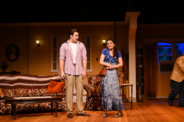 In this image from Over The River and Through The Woods, a lighthearted theatre scene in a cozy living room set. A young man in a pink plaid shirt and khakis looks at a smiling young woman in a blue patterned dress and purple top, who stands shyly with her arms crossed and a brown purse over her shoulder. The background features vintage furniture, patterned wallpaper, and warm lighting, creating a cheerful, homey atmosphere.