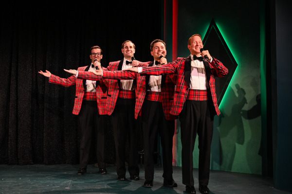 The four performers pose in a playful formation during Plaid Tidings, extending their arms outward in a showman-style gesture. They wear matching red plaid tuxedo jackets, white shirts, black trousers, and black bow ties. A dark curtain and green geometric wall lighting frame the scene.