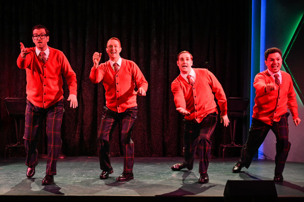 Four performers from Plaid Tidings dance energetically downstage, each reaching an arm outward toward the audience. They wear matching bright red cardigans, white shirts, plaid ties, and dark plaid trousers. The backdrop is a sparkling black curtain with two stools positioned on either side.