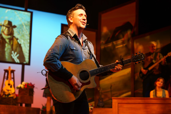 A smiling performer in a denim shirt sings and plays acoustic guitar on stage during Country Roads: The Songs of John Denver. Behind him, band members play instruments and scenic backdrops display western landscapes and a cowboy portrait. The warm lighting evokes a nostalgic, rustic atmosphere.