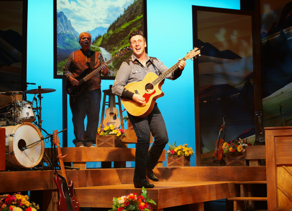 A smiling performer in a denim shirt sings and plays acoustic guitar on stage during Country Roads: The Songs of John Denver. Behind him, band members play instruments and scenic backdrops display western landscapes and a cowboy portrait. The warm lighting evokes a nostalgic, rustic atmosphere.