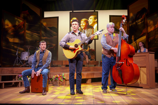 A group of performers on stage during Country Roads: The Songs of John Denver, featuring one playing guitar and singing center stage, another seated on a cajón drum, a third playing an upright bass, and a fourth at a wooden piano. The stage is decorated with warm lighting, wooden platforms, and floral arrangements, with projected vintage photos of John Denver in the background.