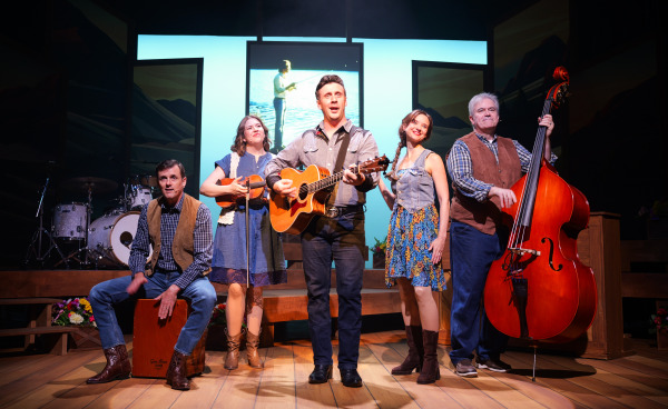 A cast of five performers on stage during Country Roads: The Songs of John Denver. The central performer sings while playing an acoustic guitar, flanked by cast members playing the cajón, violin, and upright bass, with another performer smiling nearby. Behind them, a projected image shows a person fishing in a scenic mountain lake. The stage is decorated with rustic wooden platforms and bright floral arrangements, creating a warm, nostalgic atmosphere.