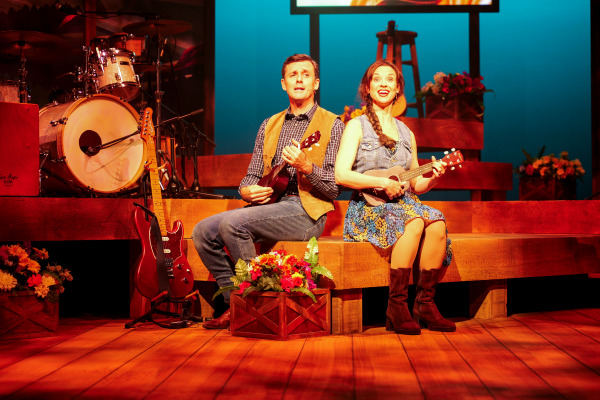 Two performers sit on a wooden stage bench during Country Roads: The Songs of John Denver, each holding a ukulele. They are brightly lit and smiling as they sing. Behind them are rustic stage elements including a drum set, a red electric guitar on a stand, floral arrangements, and a wooden stool with a guitar. The atmosphere is warm and inviting, evoking the feel of a country concert.