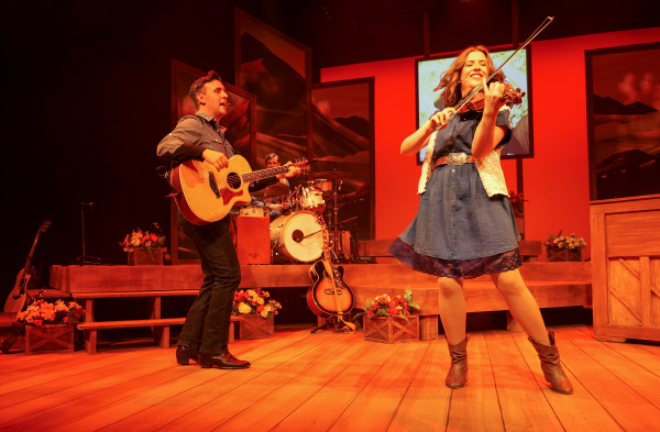 A lively musical moment from Country Roads: The Songs of John Denver, featuring a smiling fiddle player dancing at the front of the stage. A guitarist stands to the left, strumming with energy, while a drummer plays in the background. The set is decorated with warm wood textures, flower boxes, and scenic mountain artwork, all lit by a vivid orange-red glow that evokes the feel of a sunset.