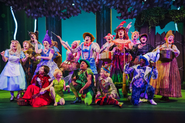 A vibrant ensemble of fairy tale creatures poses mid-song in a lively forest scene. The characters include Little Red Riding Hood, Tinker Bell, Peter Pan, Pinocchio, the Fairy Godmother, the White Rabbit, and more, all dressed in colourful, whimsical costumes with exaggerated features. They are gathered under stylized tree canopies with bright lighting and animated expressions, creating a magical and energetic atmosphere that bursts with personality and fun.