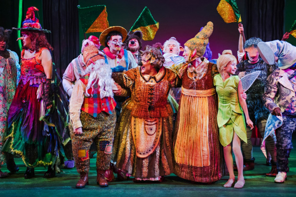 A joyful group of fairytale characters celebrates together on stage, dressed in vibrant, whimsical costumes. From left to right, characters include witches, a gnome with a long white beard, a fairy in green, and other eccentric creatures. The ensemble stands arm-in-arm, beaming with excitement, with glittering flags raised in the background and a forest backdrop behind them. The moment captures a sense of unity and playful rebellion.
