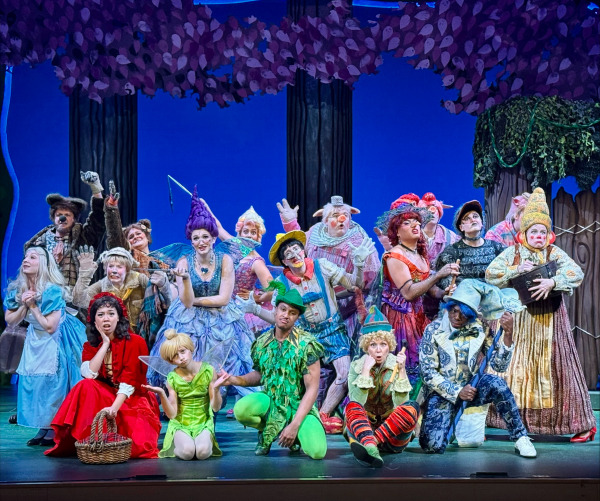 Shrek The Musical: A large ensemble of fairy-tale characters poses together onstage in a whimsical forest setting with purple treetops and a wooden fence draped in greenery. The cast includes colourful, highly stylized characters such as the Big Bad Wolf, Pinocchio, the Three Little Pigs, the Ugly Duckling, fairy-tale princesses, witches, elves, and other storybook creatures. They stand and crouch in animated poses, expressing surprise, excitement, and mischief. Bright, detailed costumes—ranging from glittering gowns to animal suits and rustic medieval attire—create a vibrant, magical tableau across the stage.