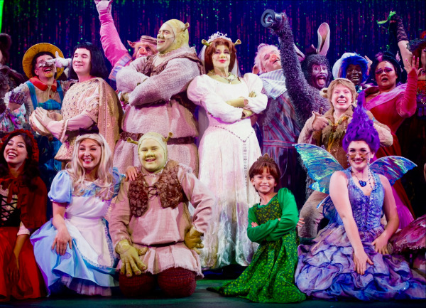 Shrek The Musical: Shrek and Fiona stand proudly at centre stage, arms crossed, surrounded by a joyful ensemble of fairy-tale characters. Shrek wears his iconic ogre costume with a padded tunic and green makeup, while Fiona appears in her white wedding gown and tiara. Around them, performers dressed as Donkey, Pinocchio, the Fairy Godmother, the Mad Hatter, a young ogre, and other storybook figures smile and pose in celebratory stances. Vibrant costumes, glittering wings, and whimsical colours fill the stage, set against a sparkling purple curtain that enhances the magical, festive atmosphere.