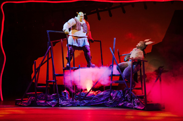 Shrek and Donkey stand atop a jagged, multi-level platform surrounded by swirling red and pink fog, simulating a dramatic and fiery setting. Shrek appears determined, gripping a railing, while Donkey lounges in a chair, looking amused or relaxed. The stage is bathed in bold red lighting, creating a sense of intensity and theatrical spectacle in this high-energy moment.
