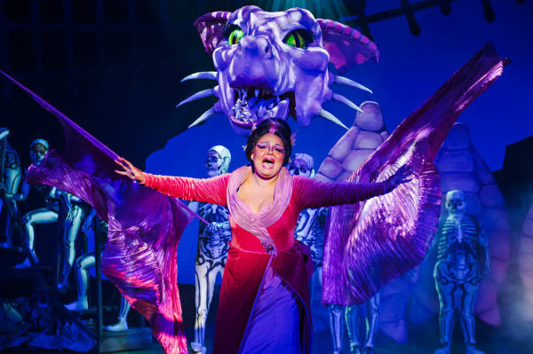 A powerful moment on stage as the Dragon, portrayed by a performer in a magenta and purple gown with shimmering wing-like sleeves, sings passionately. Behind her, a large, detailed purple dragon puppet with glowing green eyes and sharp teeth looms overhead, animated by skeletal-costumed puppeteers. The scene is lit with rich purple and pink hues, creating a dramatic, fantastical atmosphere.