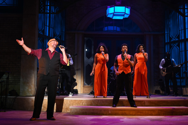 A lively group performance featuring five vocalists in coordinated red and black costumes, standing on stage with a soulful energy. On the far left, a male singer in a fedora sings expressively with one arm outstretched. Centre stage, three performers—two women in red jumpsuits and a man in a red vest—harmonize into microphones, while the band plays in the background. The set features stained glass-style windows and warm stage lighting.