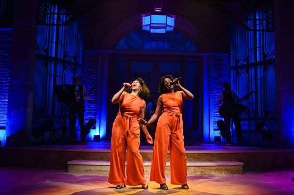 Two female singers in matching red jumpsuits stand centre stage under vivid blue lighting, holding microphones and singing passionately. They smile as they lean toward each other, creating a joyful and dynamic atmosphere. The backing band is visible in silhouette, framed by large, ornate windows.