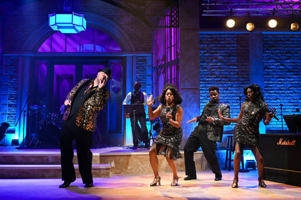 A high-energy number with four singers in glittering black and gold costumes mid-dance, striking bold poses. A male vocalist in a black and gold brocade jacket sings at the front left, while the other three—two women in fringed dresses and a man in a sequinned jacket—dance in unison. Purple and blue lighting enhance the vibrant, retro feel of the performance.