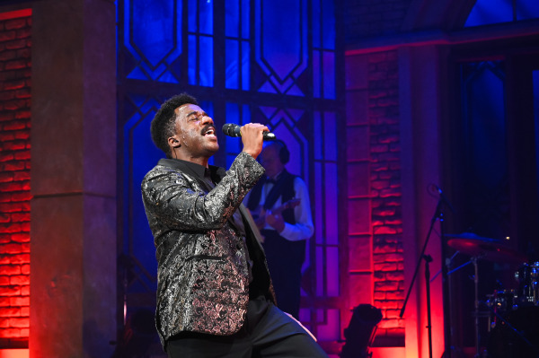 A male soloist sings with intensity, gripping the microphone and leaning forward. He wears a shimmering paisley-patterned black jacket. Behind him, the stage glows red and blue, with the band partially obscured in silhouette. The atmosphere is electric, with emotional depth conveyed in the performance.