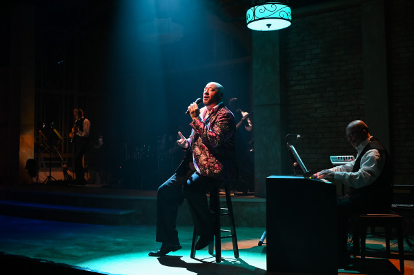  A male vocalist sits on a stool under a spotlight, singing with heartfelt expression. He wears a richly patterned jacket and holds a microphone in one hand while gesturing with the other. A pianist plays beside him under moody lighting, with other musicians visible in the shadows, creating an intimate cabaret-style moment.