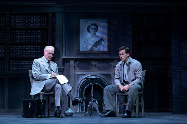 In this scene from Agatha Christie’s Witness for the Prosecution, two men sit in a dark, wood-panelled study beneath a portrait of Queen Elizabeth II. On the left, an older man in a light grey suit and glasses takes notes with a briefcase at his feet, while on the right, a younger man in a grey shirt and tie sits tensely with his hands clasped. The atmospheric lighting and detailed set evoke a formal, suspenseful courtroom drama.