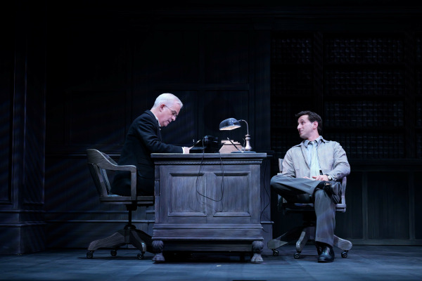 In this scene from Agatha Christie’s Witness for the Prosecution, an older man with white hair, dressed in a dark suit, sits behind a wooden desk cluttered with papers, a lamp, and a telephone, focused on his writing. Across from him, a younger man in a grey shirt and tie leans back casually in his chair, speaking with a confident expression. The dark, wood-panelled set evokes a formal office, contributing to the production’s tense, period atmosphere.