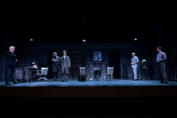 In this scene from Agatha Christie’s Witness for the Prosecution five men stand spaced across a dimly lit, wood-panelled office set, creating a tense tableau. At centre stage, two men in trench coats and fedoras appear mid-conversation, while others observe from opposite sides — one behind a desk with a lamp and telephone, another beside a judge’s wig stand. A portrait of Queen Elizabeth II hangs above a dark fireplace, adding to the play’s post-war British atmosphere and sense of intrigue.