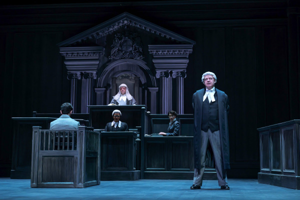A dramatic courtroom scene from Agatha Christie’s Witness for the Prosecution. The set features an imposing wooden judge’s bench framed by grand columns and the royal crest. A judge in traditional robes and wig presides from above as a barrister in a white wig and black gown addresses the court from the foreground. In front of the judge, another lawyer sits beside the witness stand, where a man in a grey suit faces forward. The dark, formal lighting heightens the intensity of the trial setting.