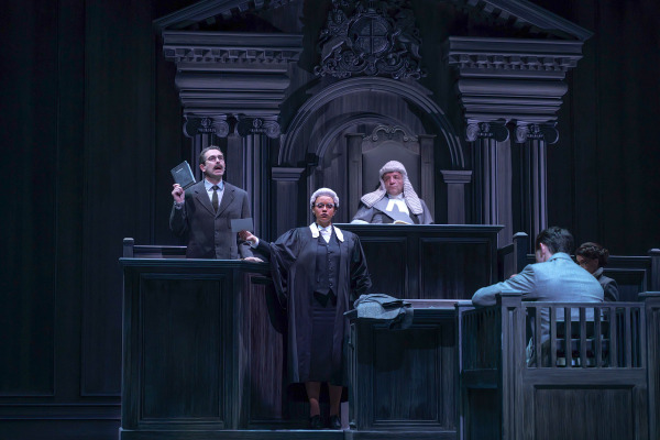 A tense courtroom moment from Agatha Christie’s Witness for the Prosecution. A man in a brown suit stands in the witness box, holding up a small book while testifying. In front of him, a barrister in traditional robes and a white wig questions him assertively, as a judge in full regalia watches from the elevated bench behind them. To the right, another man sits in the dock while a court clerk takes notes. The dramatic lighting and stately wooden set emphasize the high-stakes intensity of the trial. 