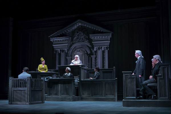 A courtroom scene from Agatha Christie’s Witness for the Prosecution. The stage depicts a grand British courtroom with wooden panelling and ornate columns framing the judge’s bench. A woman in a mustard-yellow dress and matching hat testifies from the witness stand, while the judge, wearing traditional robes and a white wig, listens attentively. Lawyers in black gowns and powdered wigs stand and sit across the stage, while the accused sits in the dock at left. The moody lighting heightens the dramatic tension of the trial.
