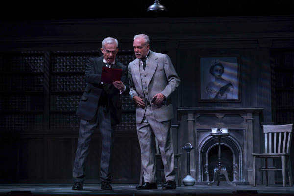 In this scene from Agatha Christie’s Witness for the Prosecution two men stand close together in a dark, wood-panelled study, both intently examining a red document folder. The man on the left, dressed in a dark suit, gestures toward the paper as he explains something urgently, while the man on the right, in a light grey three-piece suit, leans in with a serious expression. Behind them, a portrait of Queen Elizabeth II hangs above a fireplace, underscoring the play’s mid-century British setting and tone of suspense.