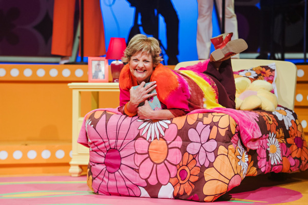 A solo performer in Yesterday Once More lies across a retro floral bedspread with a joyful expression, hugging a teal book to her chest. She wears a bright pink, orange, and yellow top with burgundy pants and red shoes. The set is awash in warm tones, with polka-dot trim, a yellow nightstand holding a small red lamp and photo frame, and hints of band members and other actors visible in the background.