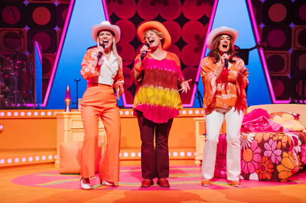 Three women stand in front of the retro bedroom set, singing with enthusiasm. Two wear white cowboy hats and one wears an orange wide-brim hat. Their orange, pink, and white costumes complement the vibrant 1970s colour palette of the set. A lava lamp and floral bed complete the playful and lively atmosphere.