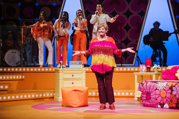   Standing center stage with open arms, a smiling older woman in a tie-dye top and maroon pants addresses the audience. Behind her, four singers in orange and white 1970s outfits sing energetically, flanked by live musicians in silhouette. The bright retro bedroom set glows with warm lighting and vintage decor.