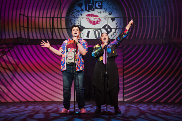 Two high school students stand centre stage, passionately singing into microphones in a vibrant, energetic performance of Mean Girls: High School Version. The student on the left wears a David Bowie T-shirt under a bright floral shirt, while the student on the right sports a paint-splattered jacket, purple tie, and black skirted pants. Behind them, a circular graphic featuring a lipstick kiss, stars, and the words “Be Kind,” “So Fetch,” and “Brave” reinforces the show's themes. A curved pink and purple striped background adds to the youthful, pop-art style atmosphere.