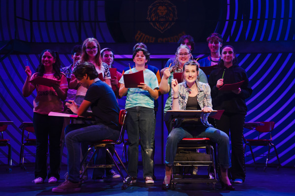 Cady Heron sits confidently at a desk in the foreground of a classroom scene, smiling and holding up a pencil, surrounded by fellow students. Behind her stands a group of teens holding red notebooks and pencils, with expressions of curiosity and excitement. A student in a black t-shirt and jeans sits at a desk beside her, looking down. The backdrop features the North Shore High School lion emblem, and purple-blue lighting with concentric stripes adds a dramatic flair to the high school setting.