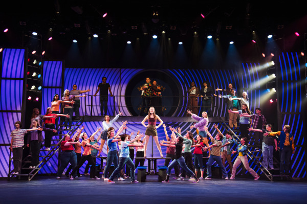A large ensemble of high school students fills the stage in a vibrant scene from Mean Girls: High School Version. A confident teen girl stands centre-stage atop a cafeteria table, arms on her hips, striking a powerful pose in a burgundy tank top and olive skirt. Students surround her on all sides, reaching out toward her with energetic choreography. Bright stage lighting and colourful costumes reflect the lively, theatrical atmosphere, while a large high school crest is visible in the background above the second tier of the multi-level set.