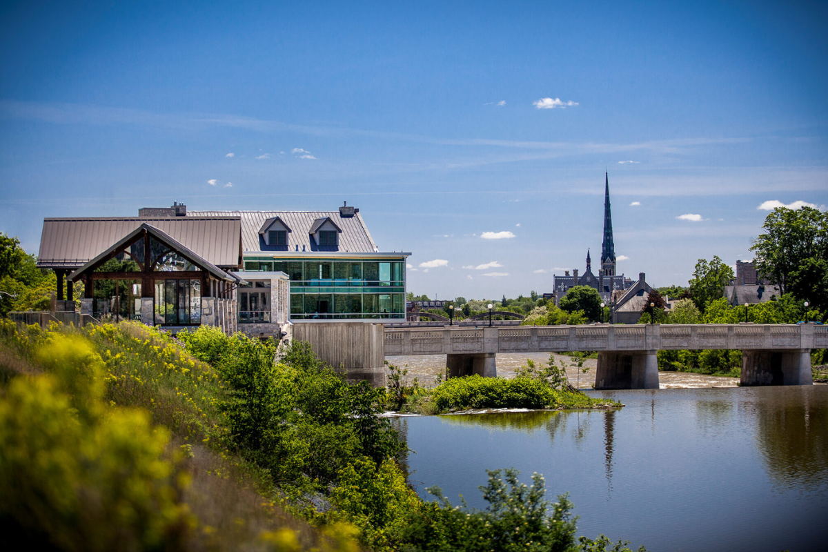 This image captures a scenic outdoor view with a modern building featuring large glass windows on the left, overlooking a river. A stone bridge spans the river, leading to a cityscape with a prominent church steeple in the distance, all under a brigh