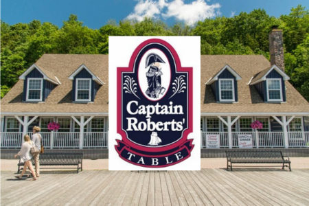 A waterfront building with tan shingles, white trim, and two dormer windows on each side, framed by lush green trees and a blue sky. In front of the building, two benches face a wide wooden boardwalk where a couple walks hand in hand. Superimposed in