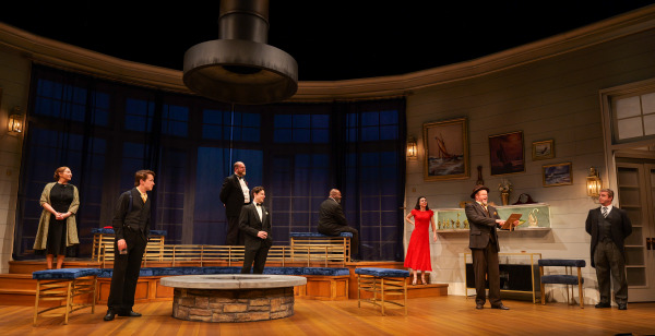 A formal gathering takes place in Agatha Christie’s And Then There Were None as several performers stand and sit across a spacious modern interior set with curved walls, blue-lit windows, and tiered platforms. One man in a tan suit and fedora reads dramatically from a piece of paper while the surrounding characters watch intently. A woman in a bright red dress stands near the mantelpiece, adding contrast to the otherwise muted palette of the sophisticated set.