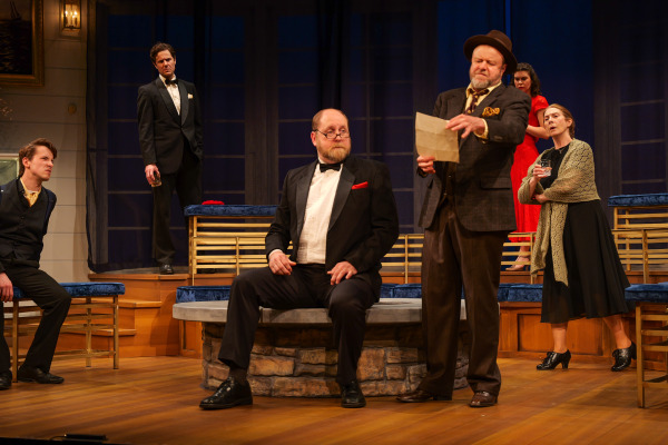 A tense ensemble scene unfolds in Agatha Christie’s And Then There Were None as a man in a tan suit and fedora dramatically reads from a sheet of paper beside a seated man in a black tuxedo with a red pocket square. Around them, performers react with concern and suspicion from different levels of the tiered set, while warm theatrical lighting enhances the atmosphere of mystery and unease.