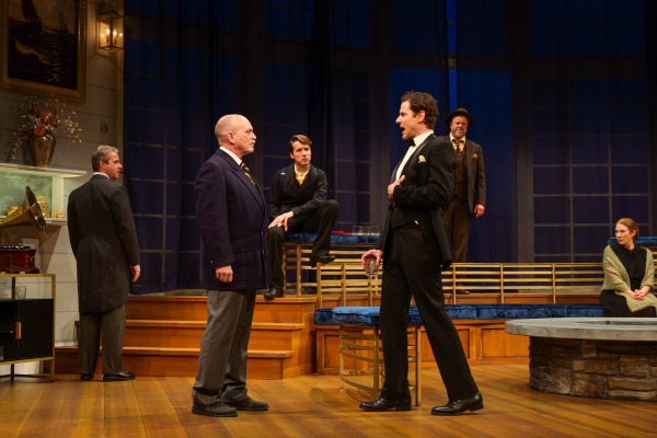 A heated confrontation takes centre stage in Agatha Christie’s And Then There Were None as two men face each other in the middle of an elegant coastal home interior. One performer in a black tuxedo gestures passionately while holding a drink, while the older man opposite him stands firmly in place. Other characters observe silently from the background, increasing the dramatic tension of the scene.