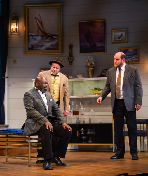 A suspenseful moment unfolds in Agatha Christie’s And Then There Were None as a seated man in a plaid jacket turns sharply toward something offstage while two men behind him look on with concern. The scene takes place within a refined seaside home set featuring nautical paintings, glass shelving, and layered wooden platforms illuminated by warm lighting.