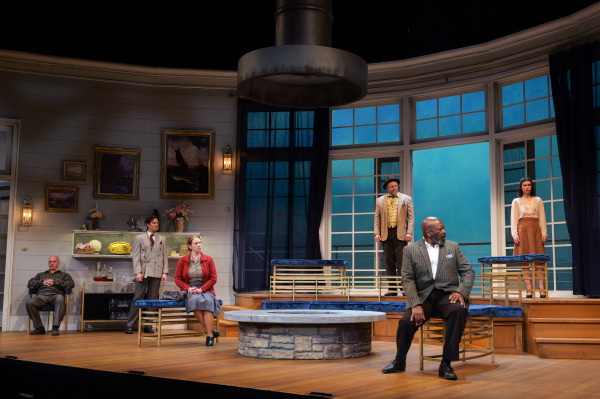 A wide ensemble tableau from Agatha Christie’s And Then There Were None shows several performers positioned throughout a modern coastal home set with tall windows and curved walls. One man sits tensely at centre stage while other characters stand or sit around him at varying heights on the tiered platforms. Cool blue light filters through the windows, contrasting with the warm tones of the interior and emphasizing the suspenseful mood.