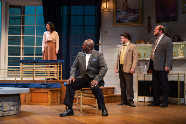A dramatic scene unfolds in Agatha Christie’s And Then There Were None as a seated man in a plaid jacket looks sharply toward a woman standing on a raised platform near tall blue-lit windows. Two men stand nearby watching intently, including one in a tan jacket and fedora and another in a grey suit. Nautical artwork, glass shelving, and layered wooden platforms frame the elegant yet tense atmosphere of the scene.