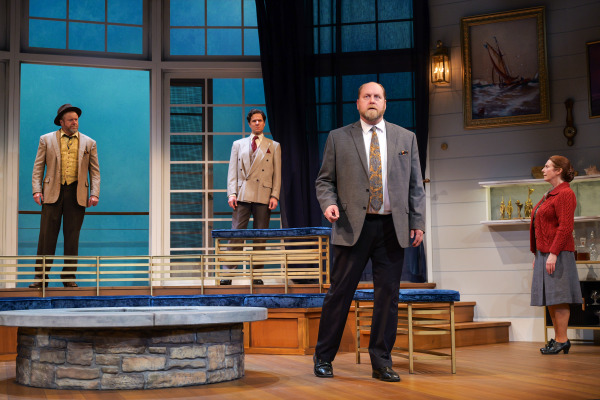 A dramatic moment unfolds in Agatha Christie’s And Then There Were None as four performers stand spread across a modern coastal home set with tall blue-lit windows and tiered wooden platforms. A man in a grey suit stands downstage centre with a tense expression while two men behind him watch silently from an upper level. Off to the side, a woman in a red cardigan and grey skirt stands near nautical paintings and glass shelving, adding to the suspenseful atmosphere.