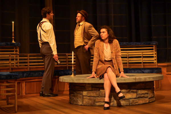 A tense exchange takes place in Agatha Christie’s And Then There Were None as three performers gather around a circular stone structure at centre stage. A young man in suspenders faces an older man in a tan jacket and fedora while a woman in a cardigan and skirt sits nearby with a wary expression. Candles and warm amber lighting cast dramatic shadows across the elegant wooden set.