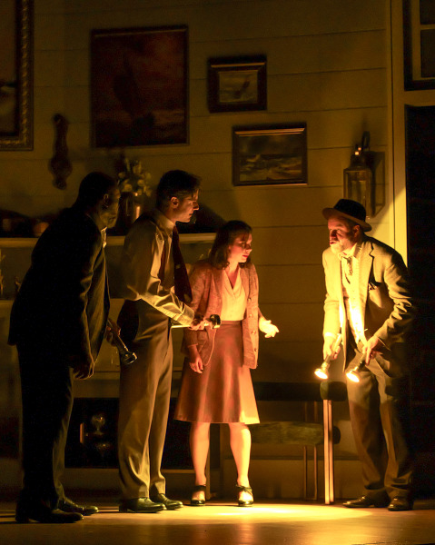 A suspenseful investigation scene unfolds in Agatha Christie’s And Then There Were None as four performers cautiously search through a darkened room using flashlights. The beams of light illuminate worried expressions and cast long shadows across the stage, while nautical paintings and dim wall sconces heighten the mysterious mood of the coastal home interior.